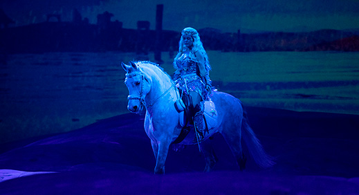 Vaaleatukkainen nainen lavalla valkoisen hevosen selässä