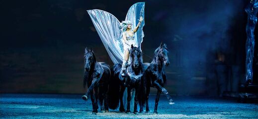 Giulia Giona als Amazone des Windes reitet stehend auf vier Friesen