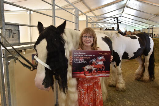 Mädchen steht vor Tinker Pferd und hält Gutschein für Reittherapie hoch